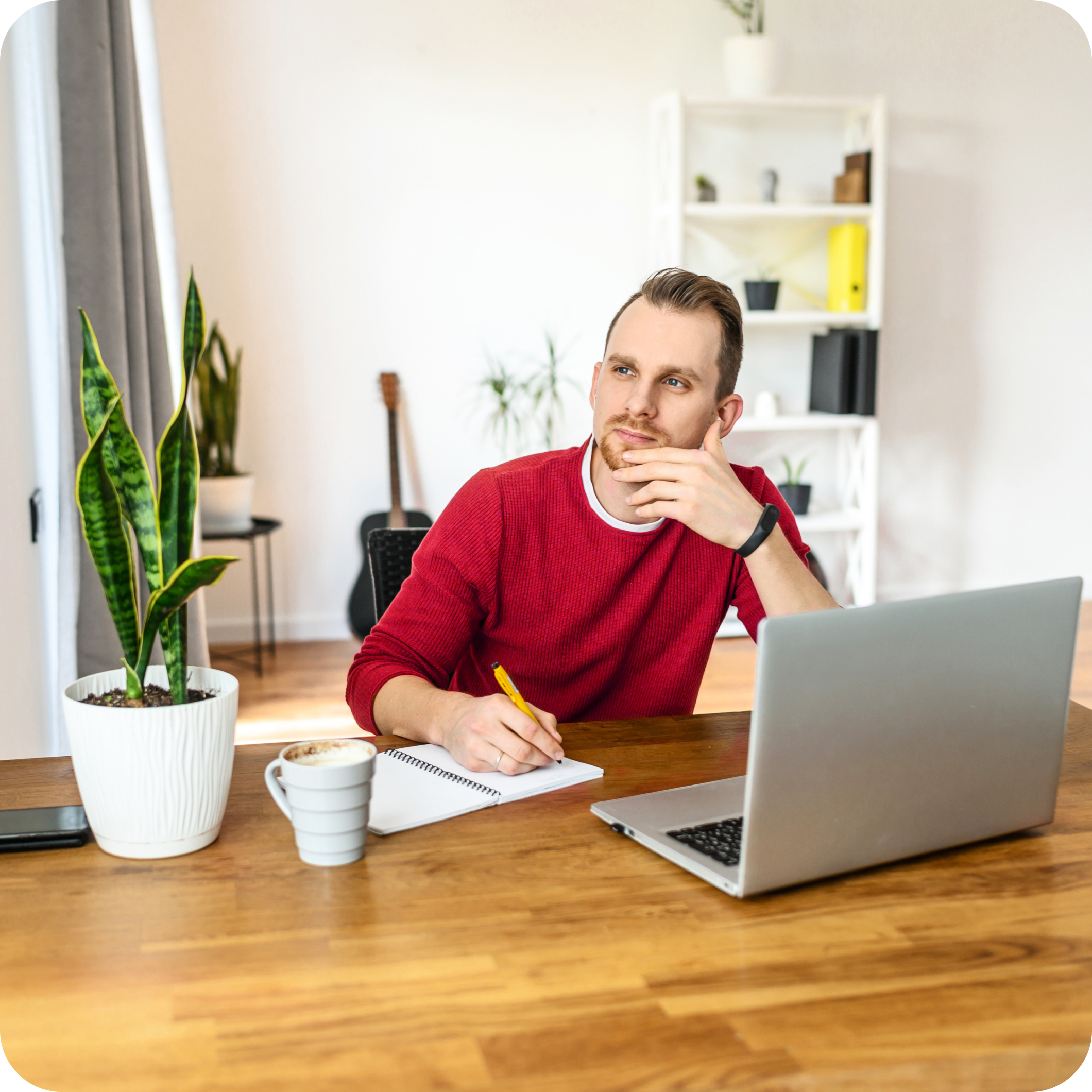 Young man with goatee and red sweater sitting at desk in a thoughtful pose, thinking what he wants to write for his online dating profile on his paper or his laptop.
