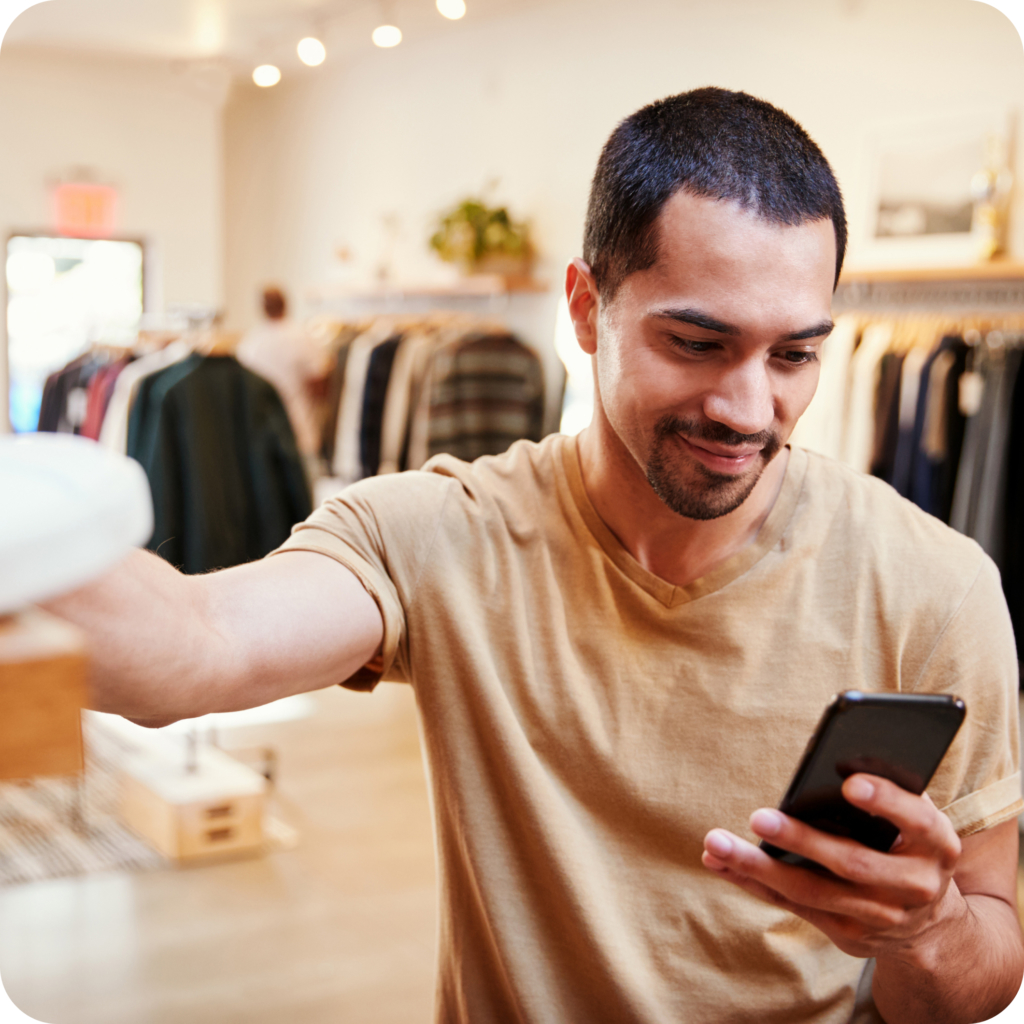 young hispanic man in light tan t-shirt looking at his cell phone reviewing wardrobe styling selections at clothing store